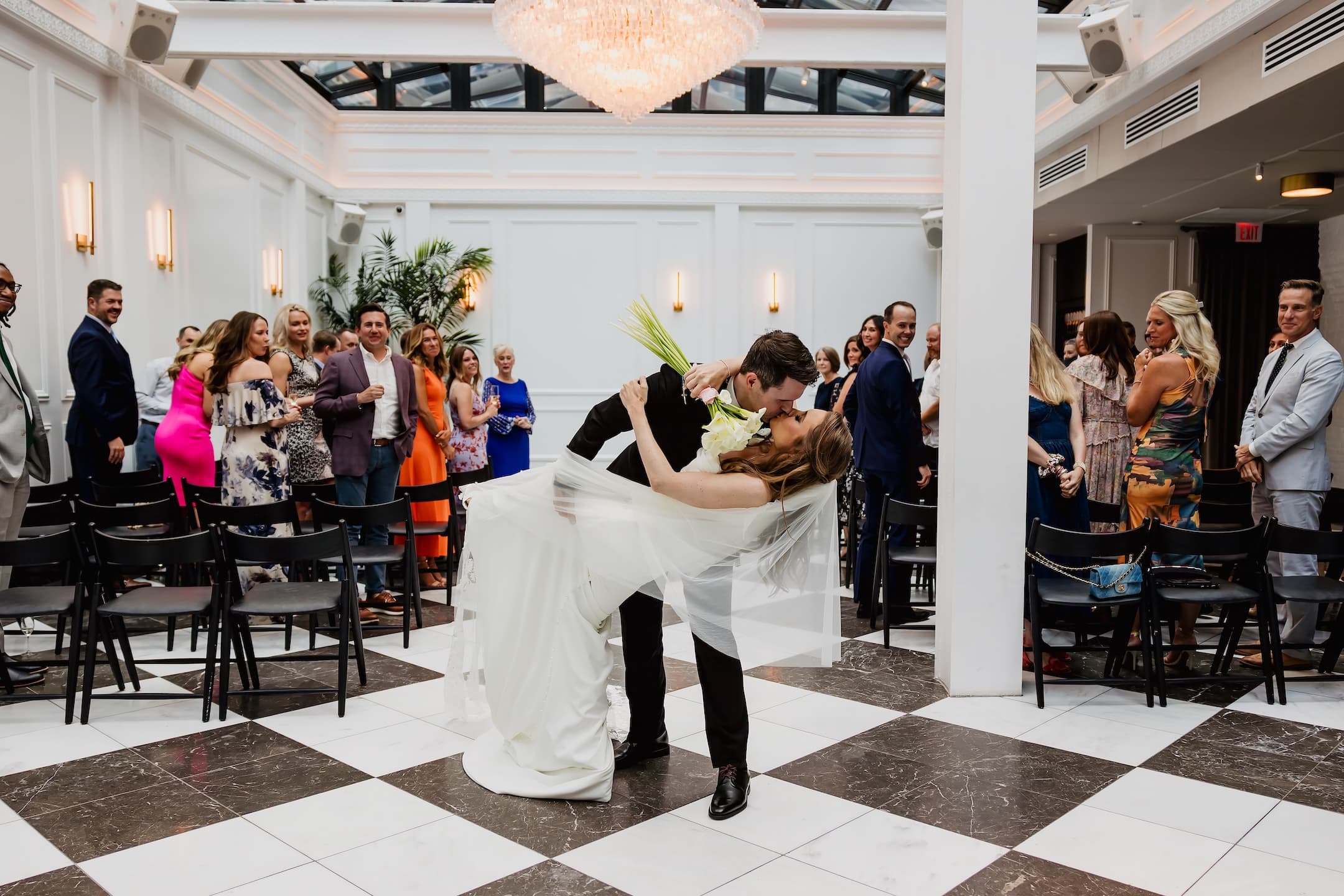 A bride and group kissing and dipping back under a chandelier with guests on either side of the aisle looking back at them and smiling.
