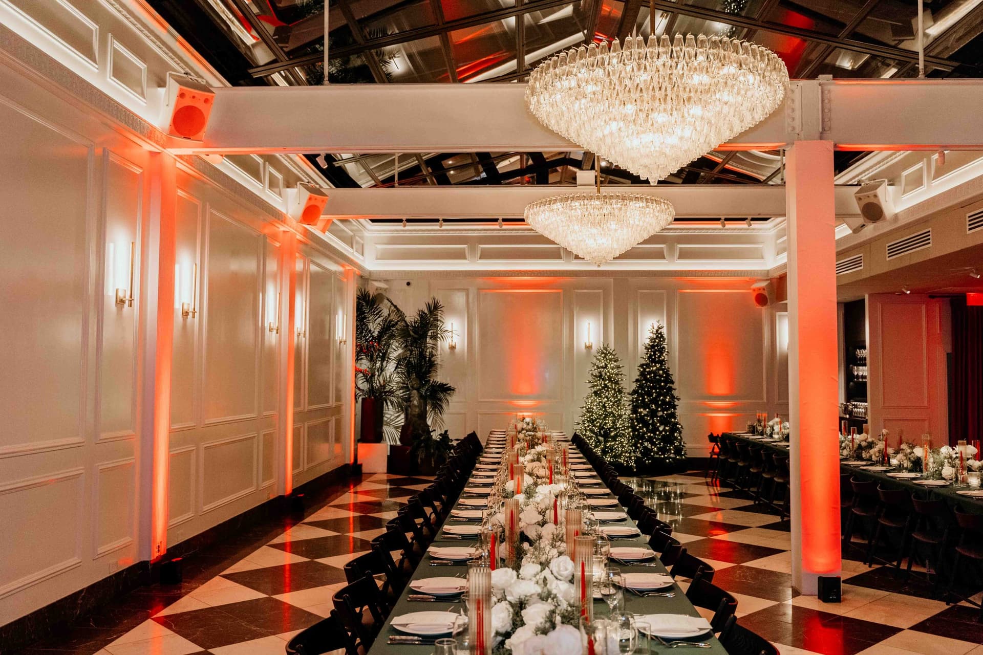 A long table in a room with a checkered floor and a skylight ceiling with chandeliers hanging. Red lights illuminate the space and flowers decorate the table.